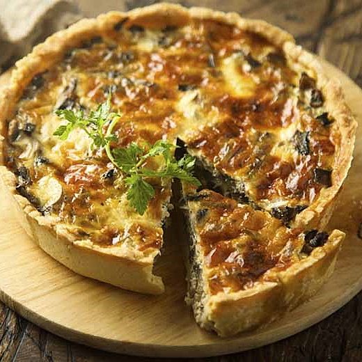Une tarte poireau-chèvre, posé sur un plateau en bois. Elle illustre la recette poireau-chèvre de La ferme du Paragard. Dessus se trouve une feuille de persil. Une part est coupé.
